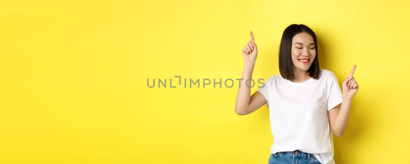 Happy asian woman dancing and having fun, posing in white t-shirt against yellow background.