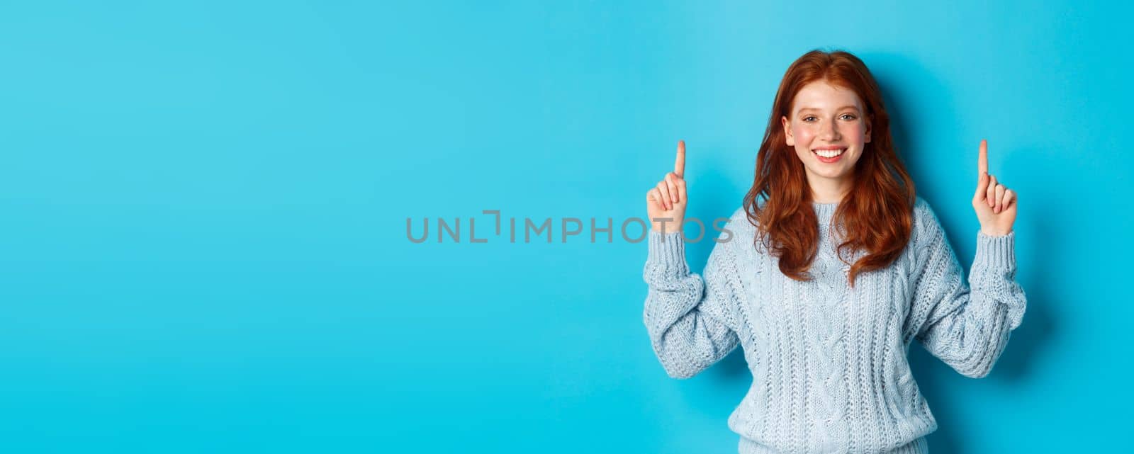 Winter holidays and people concept. Cheerful redhead girl in sweater pointing fingers up, showing logo banner and smiling, standing over blue background.