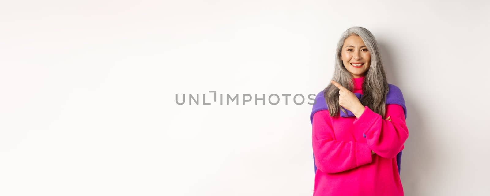 Portrait of happy asian senior woman smiling, pointing at upper left corner and looking satisfied, demonstrate special promotion, standing over white background.