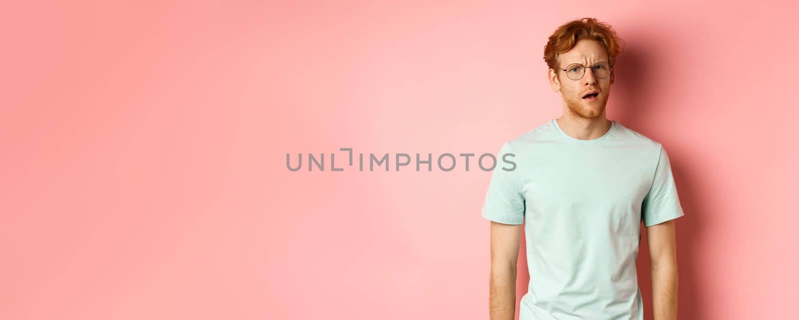 Portrait of handsome redhead man in glasses looking confused, open mouth and stare at camera, cant understand nothing, standing over pink background.