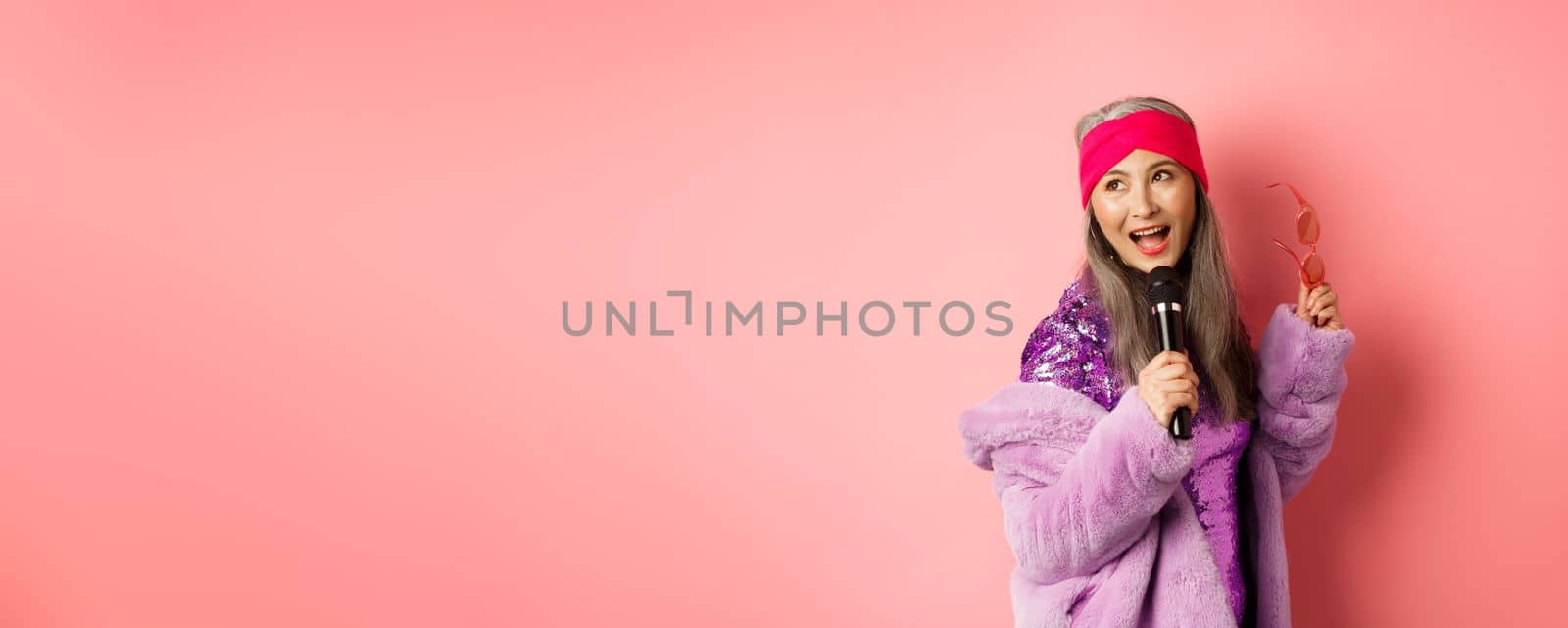 Gorgeous asian senior woman singing karaoke in mic, perform song and looking happy, standing over pink background.