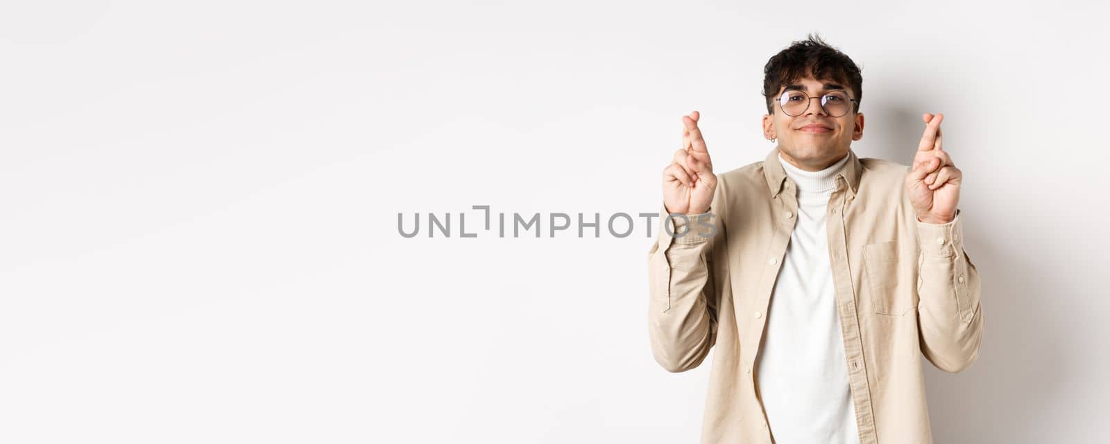 Hope. Happy young man making wish and smiling, looking hopeful at camera as waiting for dream come true, cross fingers for good luck, standing on white background.