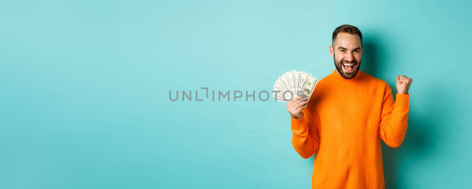 Shopping. Cheerful guy holding money, winning prize in cash and making fist pump, triumphing with satisfied expression, standing in orange sweater against turquoise background.