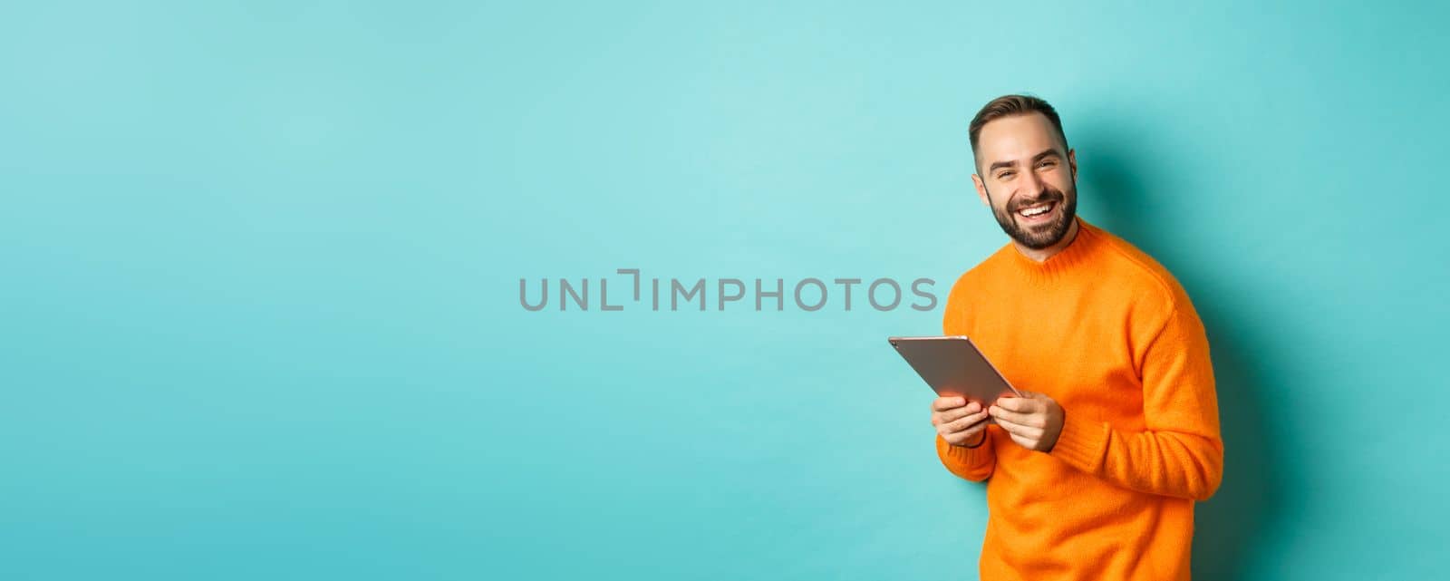 Handsome bearded man using digital tablet, laughing at camera, standing happy against light blue background.