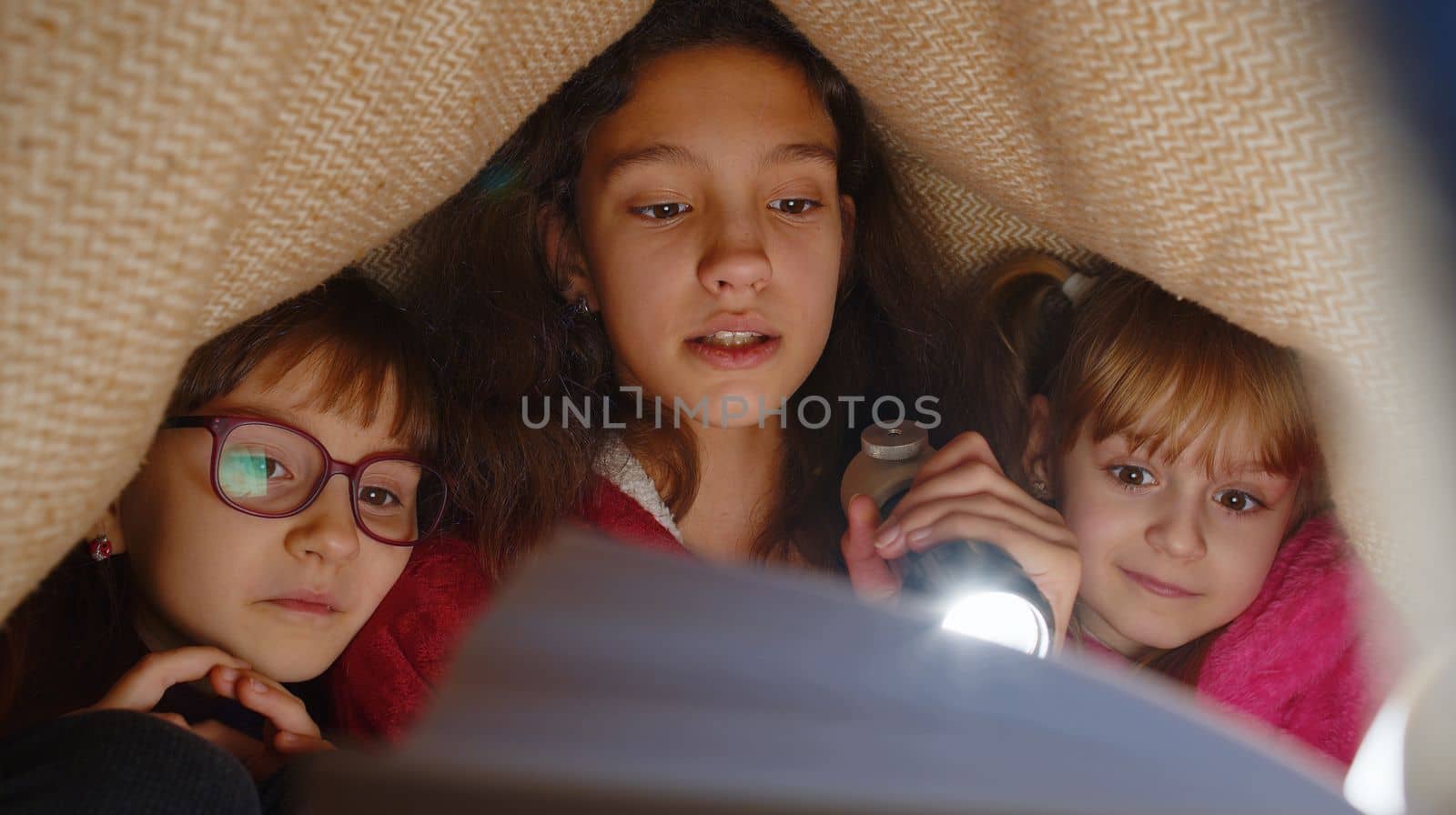 Teen girl and toddler child sisters under blanket plaid covers with flashlight reading interesting fairytale story book using flashlight. Female three siblings children kids friends in wigwam at home