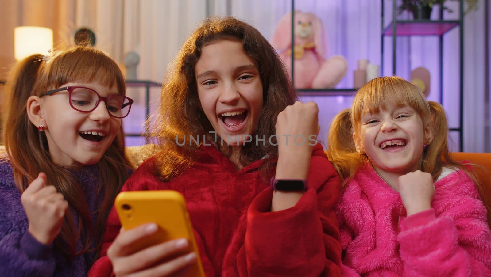 Teenage girl and little toddler child sisters using playing on mobile smartphone typing browsing say Wow yes found out great big game win good news celebrate. Three siblings children kids at home room