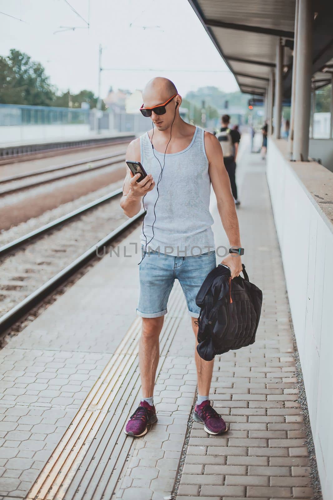 Young man in a t-shirt on the platform waiting for a train using mobile phone. Man by train station platform suburban with bag and mobile phone. Traveling by train