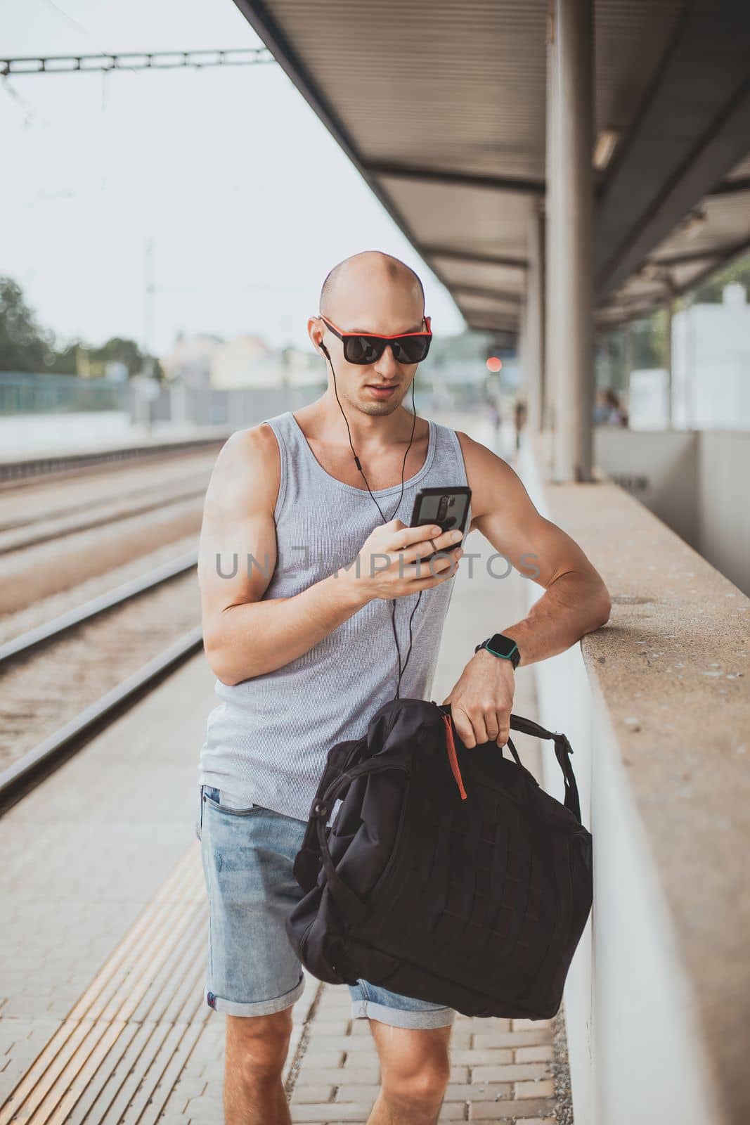 Young man in a t-shirt on the platform waiting for a train using mobile phone. Man by train station platform suburban with bag and mobile phone. Traveling by train