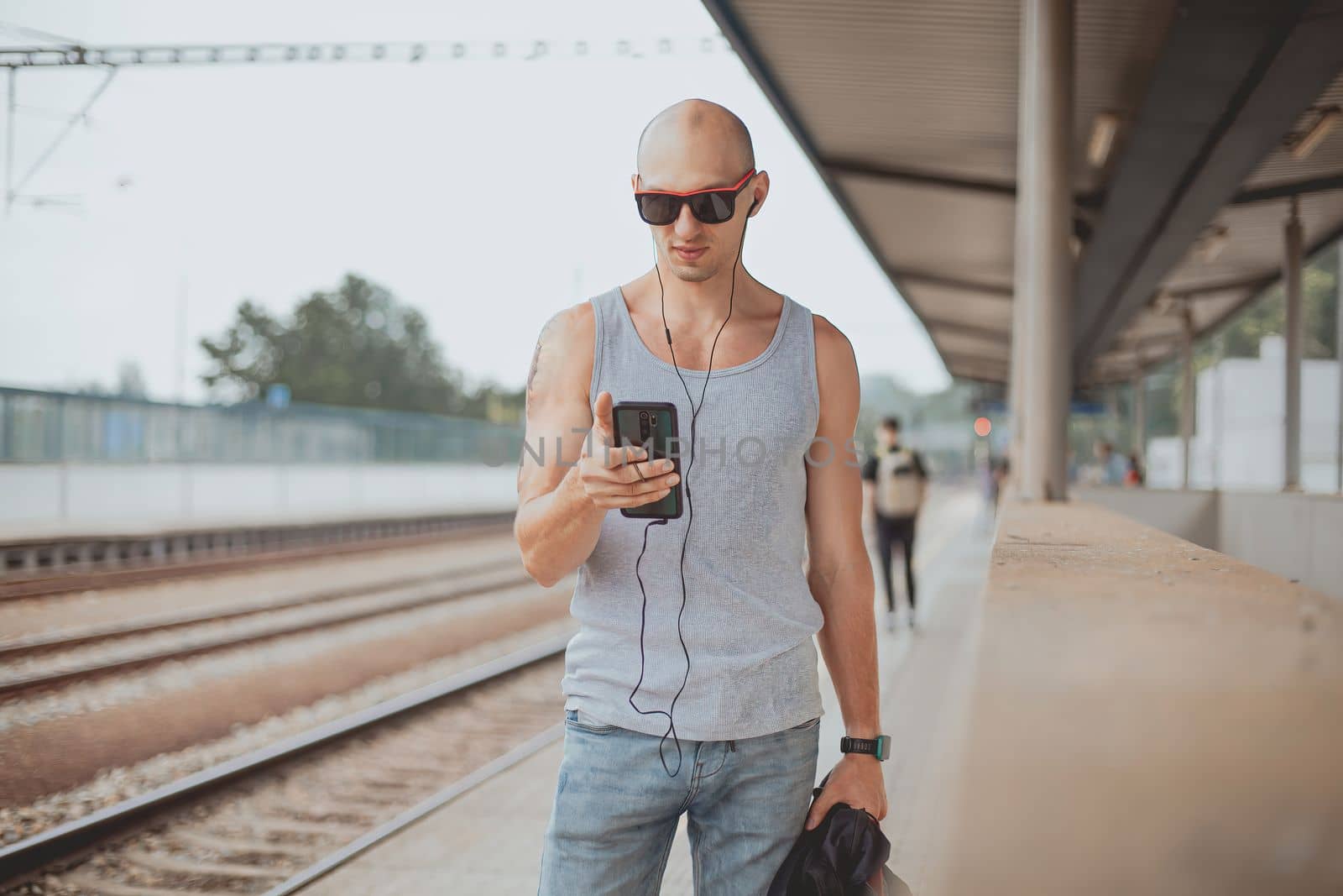 Young man in a t-shirt on the platform waiting for a train using mobile phone. Man by train station platform suburban with bag and mobile phone. Traveling by train