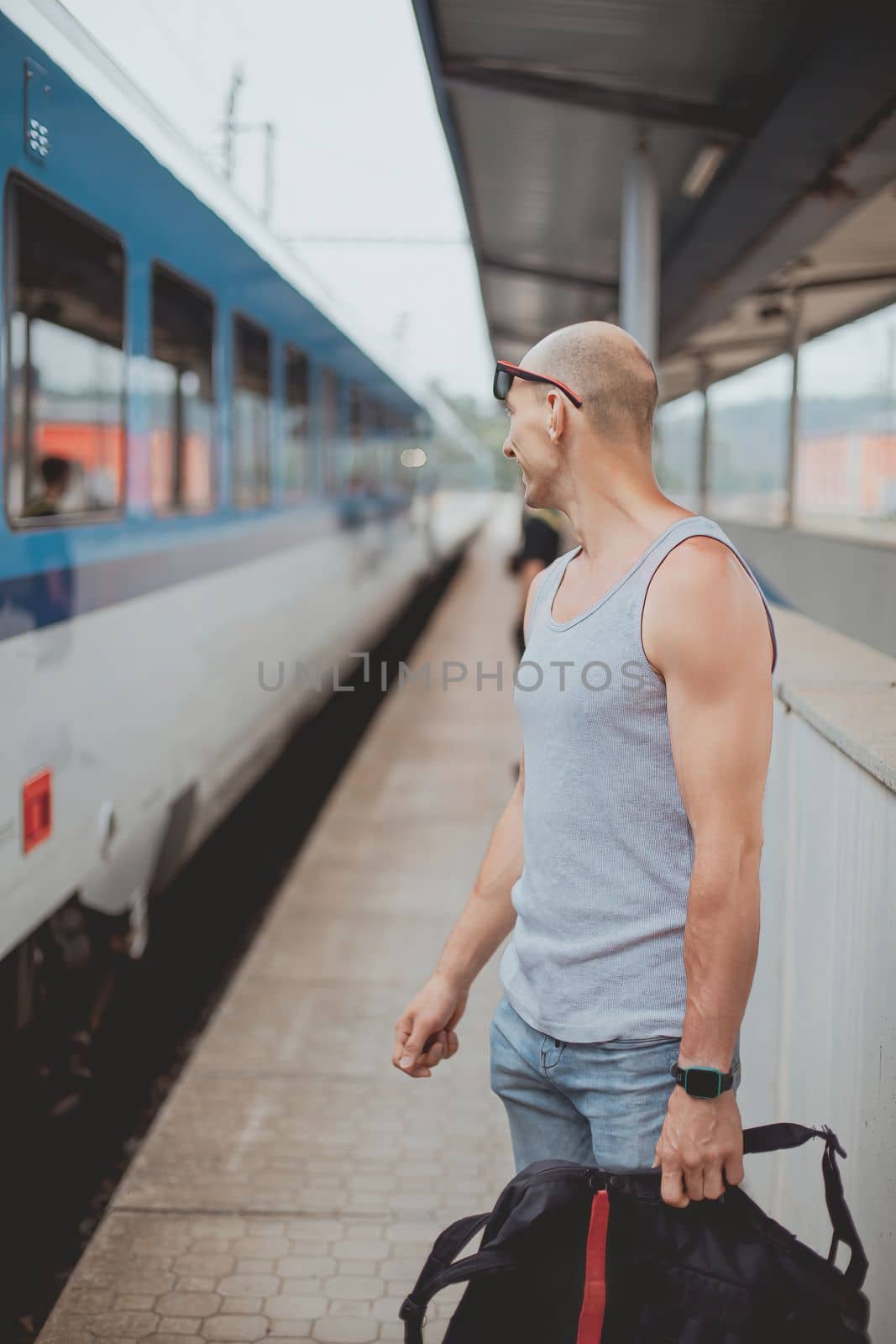 Young man in a t-shirt on the platform waiting for a train using mobile phone. Man by train station platform suburban with bag and mobile phone. Traveling by train