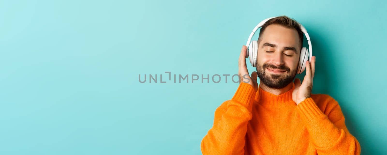 Close-up of handsome modern man listening music in headphones, standing in orange sweater over turquoise background.