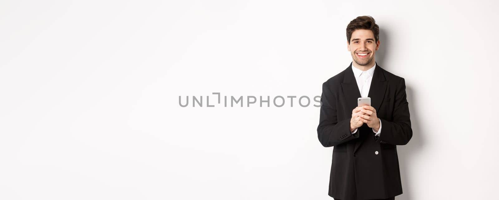 Portrait of successful, handsome businessman in suit, writing message on smartphone and smiling, standing over white background.