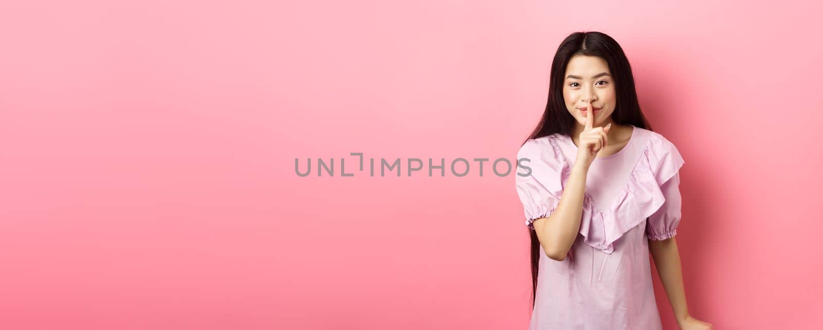 Cute asian girl hiding secret, hushing with finger pressed to lips and smiling, asking to keep quiet, standing in dress on pink background by Benzoix