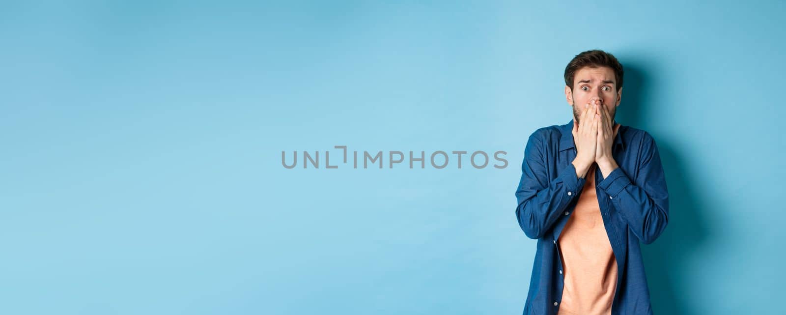 Scared anxious man covering mouth with hands and looking at something terrifying, standing on blue background.