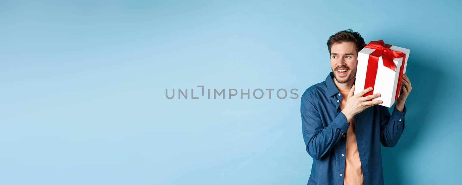 Valentines day. Smiling man guessing what inside gift box, got present from lover girlfriend, standing against blue background. Copy space