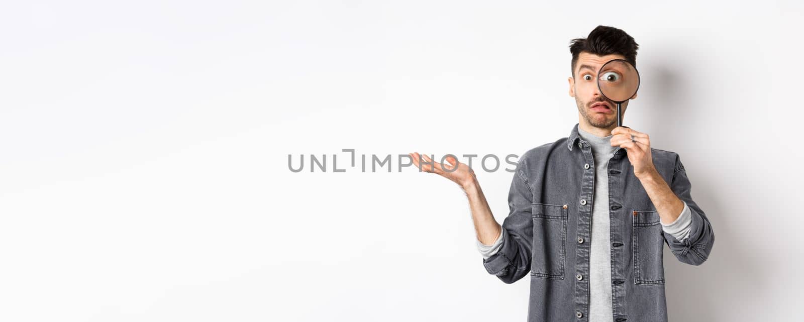 Confused man look through magnifying glass and shrugging clueless, standing on white background by Benzoix