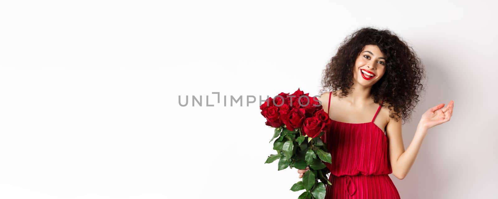 Happy woman celebrating, wearing stylish dress and holding flowers, smiling at camera, white background.