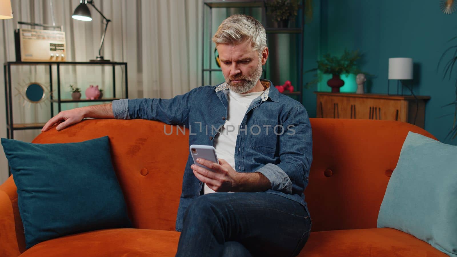 Bearded middle-aged man sitting on sofa uses mobile phone smiles at night home apartment. Mature guy texting share messages content on smartphone social media applications online, watching relax movie