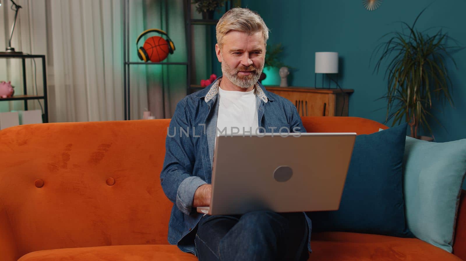 Handsome middle-aged man freelancer at home night room sitting on couch, opens laptop start working. Mature old guy works on notebook, sends messages, makes online purchases, watching movies, working