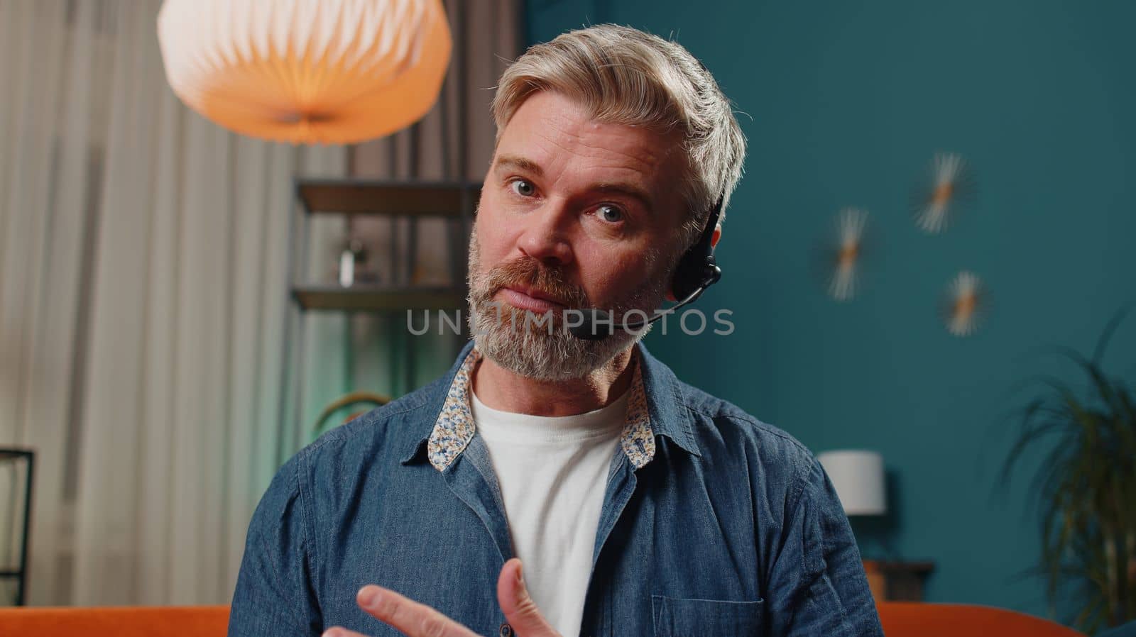POV of senior man at home wearing headset, freelance worker, call center or support service operator helpline, having talk with client or colleague communication support. Mature guy in evening room