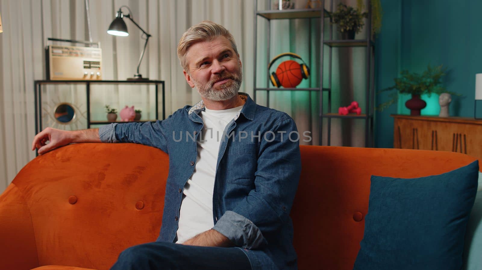 Happy calm middle-aged senior man at home couch smiling friendly, glad expression looking away dreaming resting, relaxation feel satisfied concept good news, celebrate win. Mature guy in night room