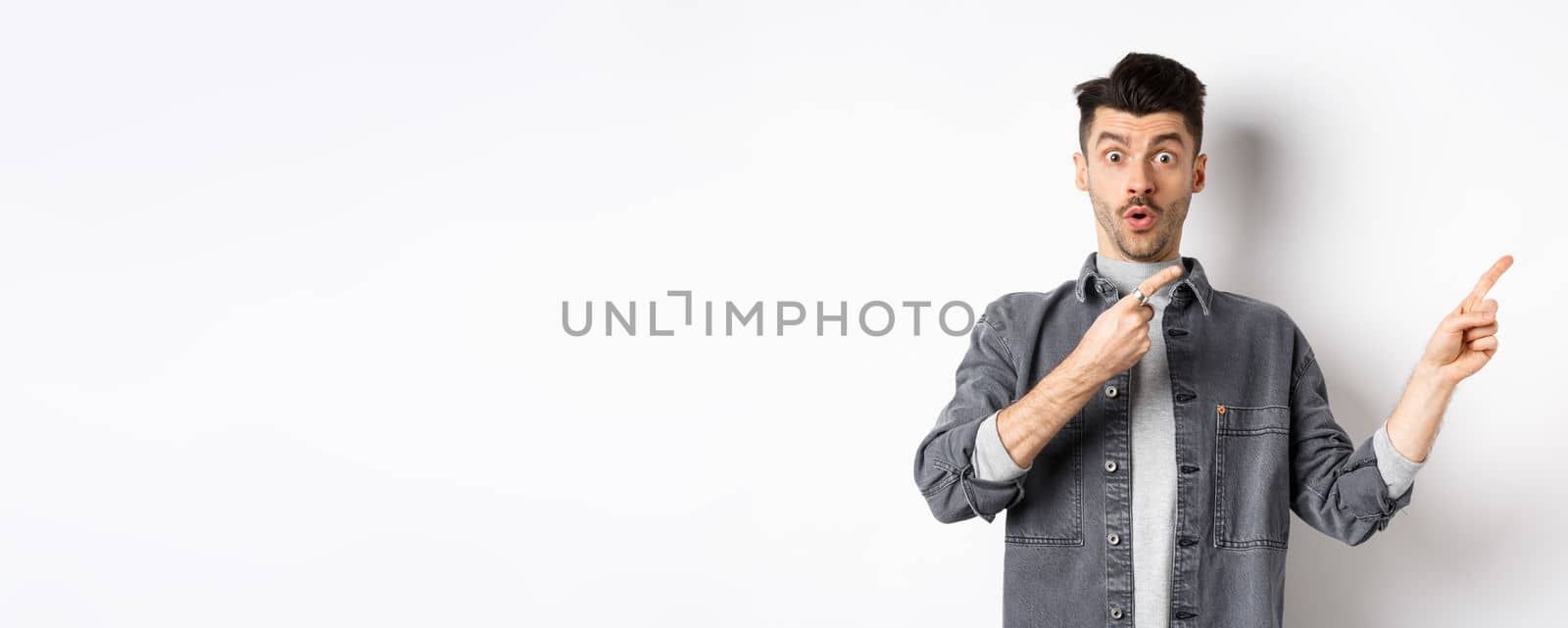 Excited guy showing advertisement aside, pointing fingers right and gasping amazed, checking out special deal, standing on white background.
