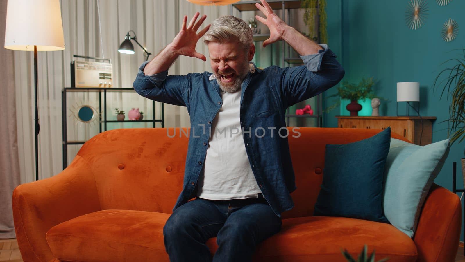 Scared mature senior man screams from stress tension problems panic horror hopelessness fear surprise shock expresses gestures rage. Furious middle-aged guy at home in room on orange couch. Lifestyle