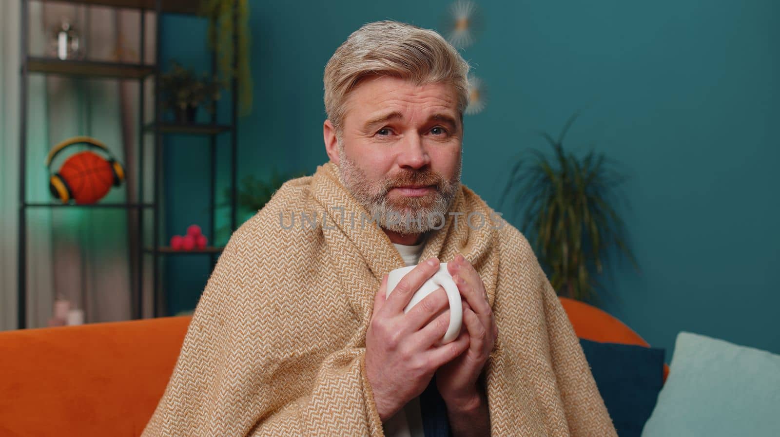 Sick man wrapped in plaid sit alone shivering from cold on sofa drinking hot tea in unheated apartment without heating due debt. Unhealthy mature guy feeling discomfort try to warm up at evening home