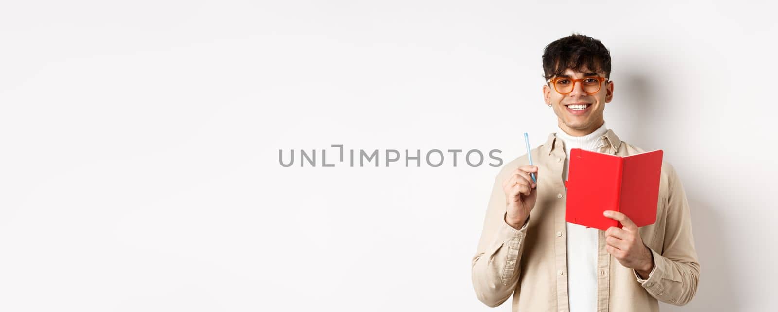 Happy person in glasses writing in journal, holding pen and diary, smiling at camera, plan a schedule, standing with planner on white background.