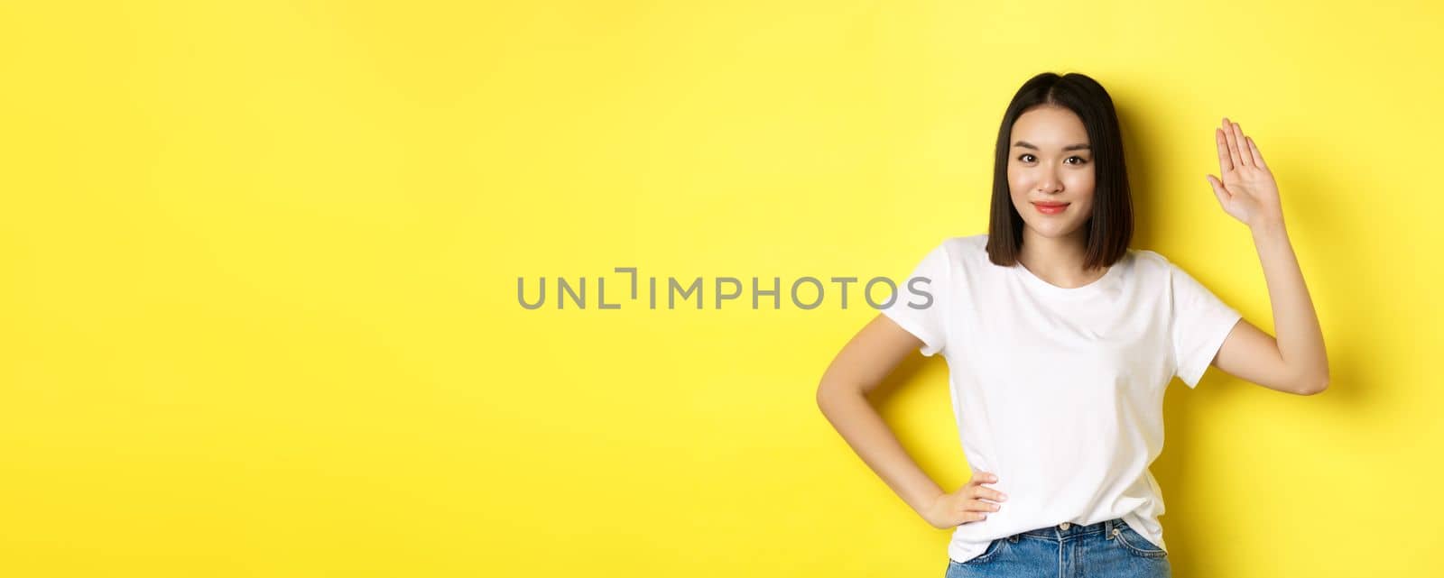 Friendly asian woman in white t-shirt waving hand and saying hello, greeting you, standing over yellow background by Benzoix