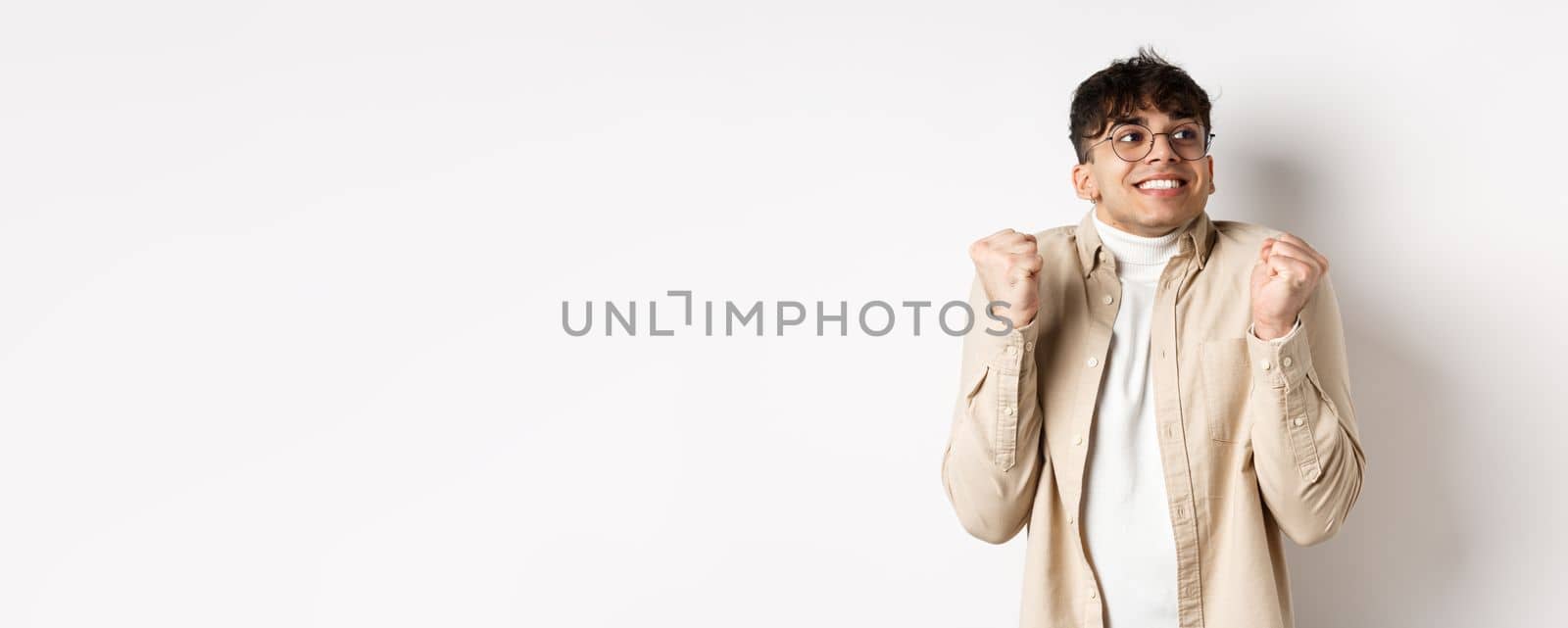 Image of handsome excited man feeling motivated and lucky, looking right and smiling, making fist pump gesture to celebrate victory, winning prize, standing on white background.