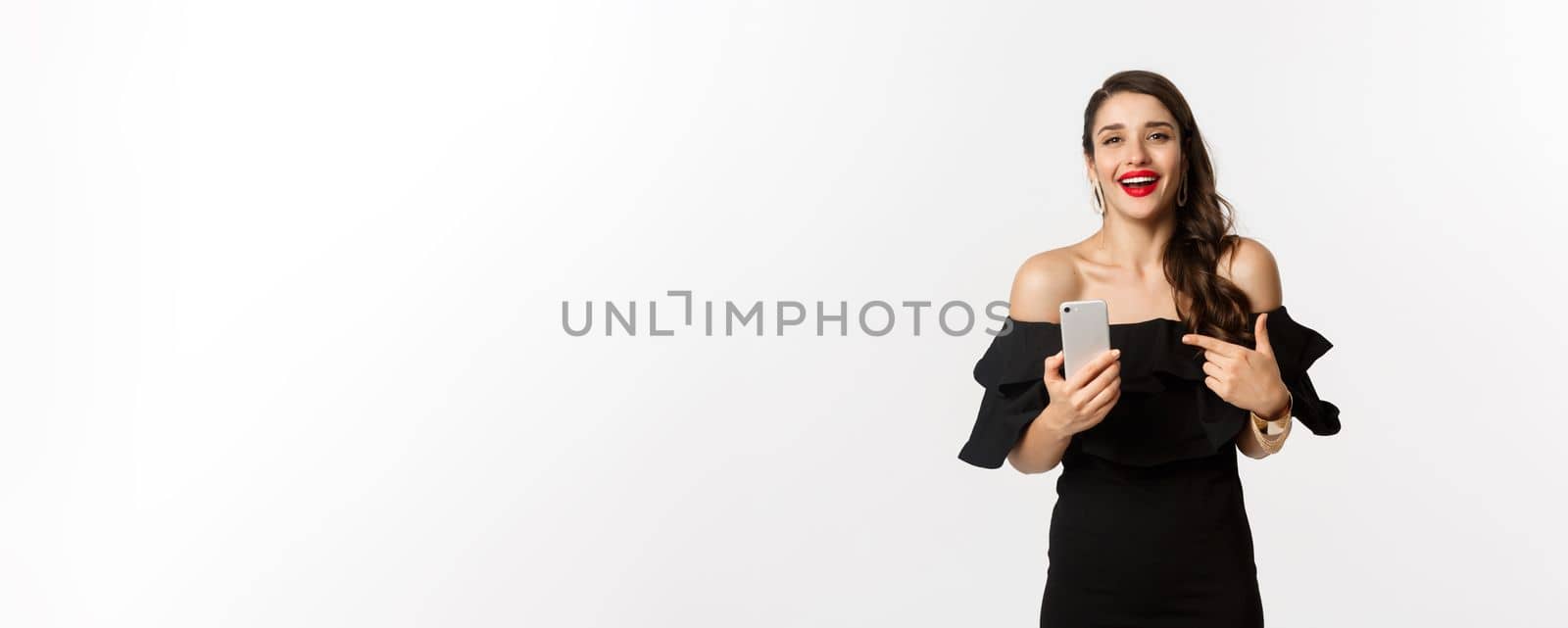 Online shopping concept. Satisfied pretty woman in black dress, smiling pleased and pointing at mobile phone, standing over white background.