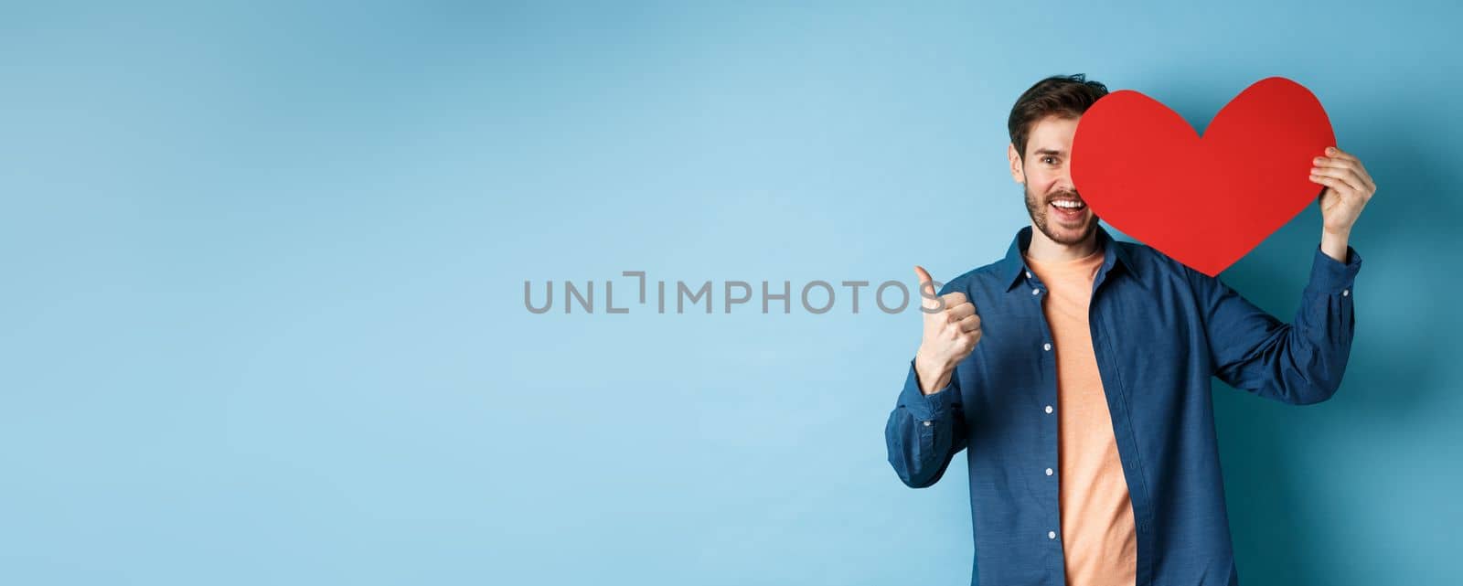 Happy man showing valentines heart and thumbs-up gesture, standing over blue background by Benzoix