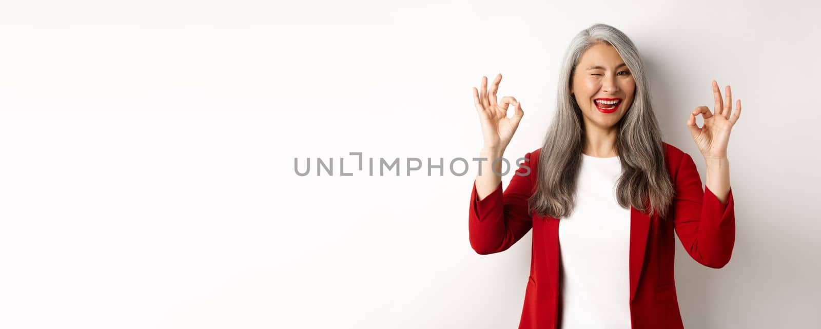 Asian professional businesswoman showing OK signs and winking, smiling pleased, assure or recommend something, standing against white background.
