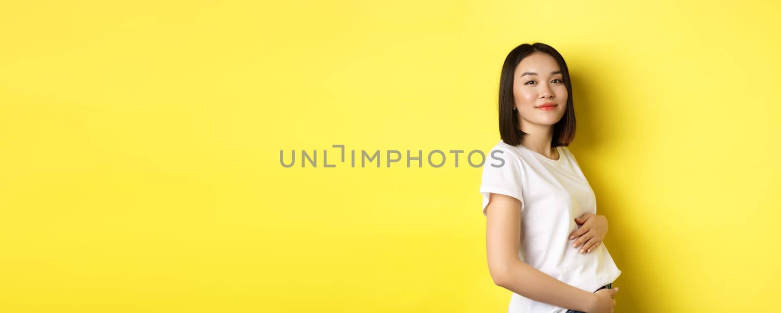 Asian woman smiling kindly, touching belly and looking at camera, being pregnant, having a baby, standing over yellow background by Benzoix