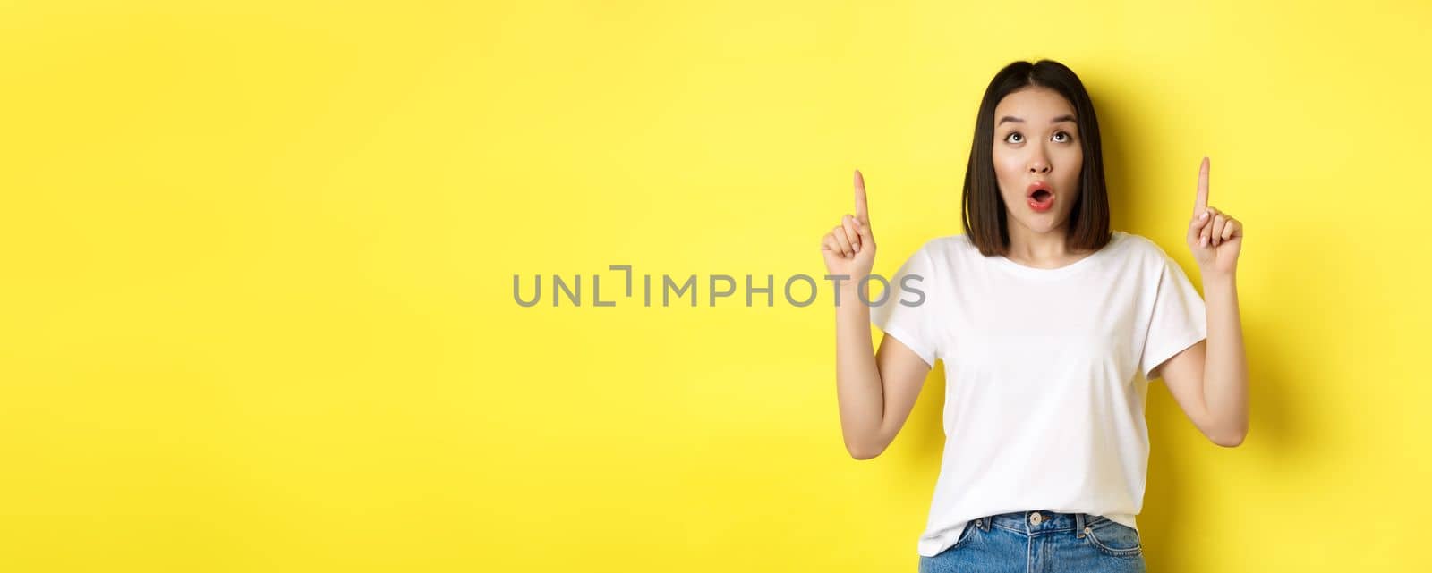 Beauty and fashion concept. Beautiful asian woman in white t-shirt pointing fingers up, standing over yellow background by Benzoix