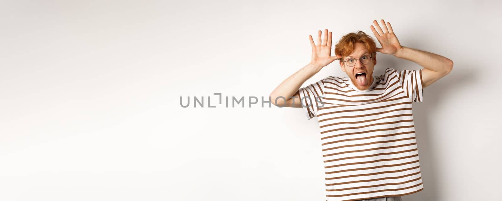 Funny young redhead man acting silly, showing tongue and mocking someone with stupid face, standing over white background.