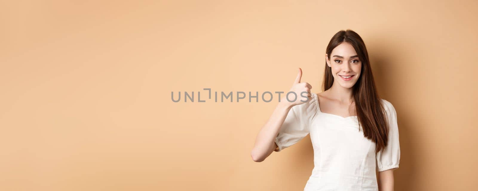 Good job. Smiling candid woman in blouse showing thumb up, praise nice choice, recommending product, standing satisfied on beige background.
