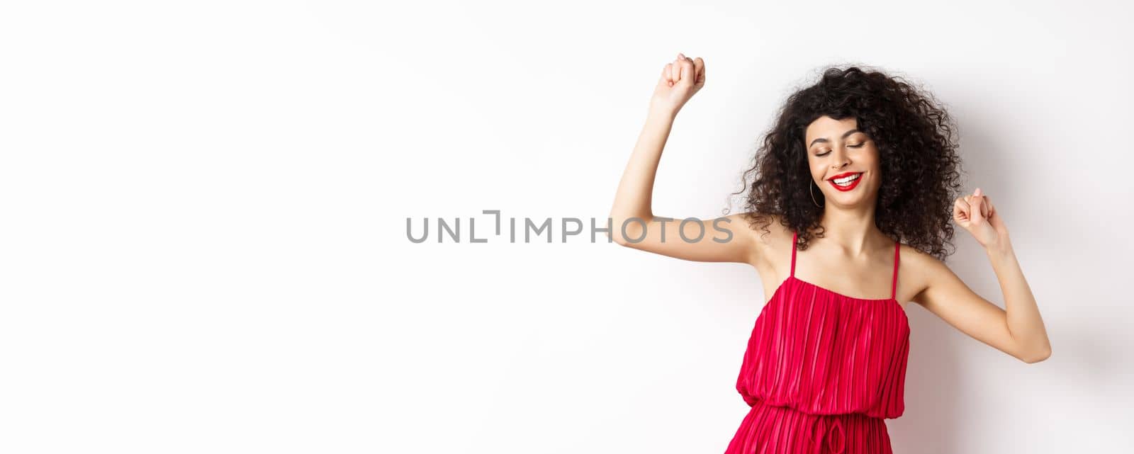 Carefree woman with curly hair, dancing at party, wearing red dress, relaxing with music, standing on white background by Benzoix