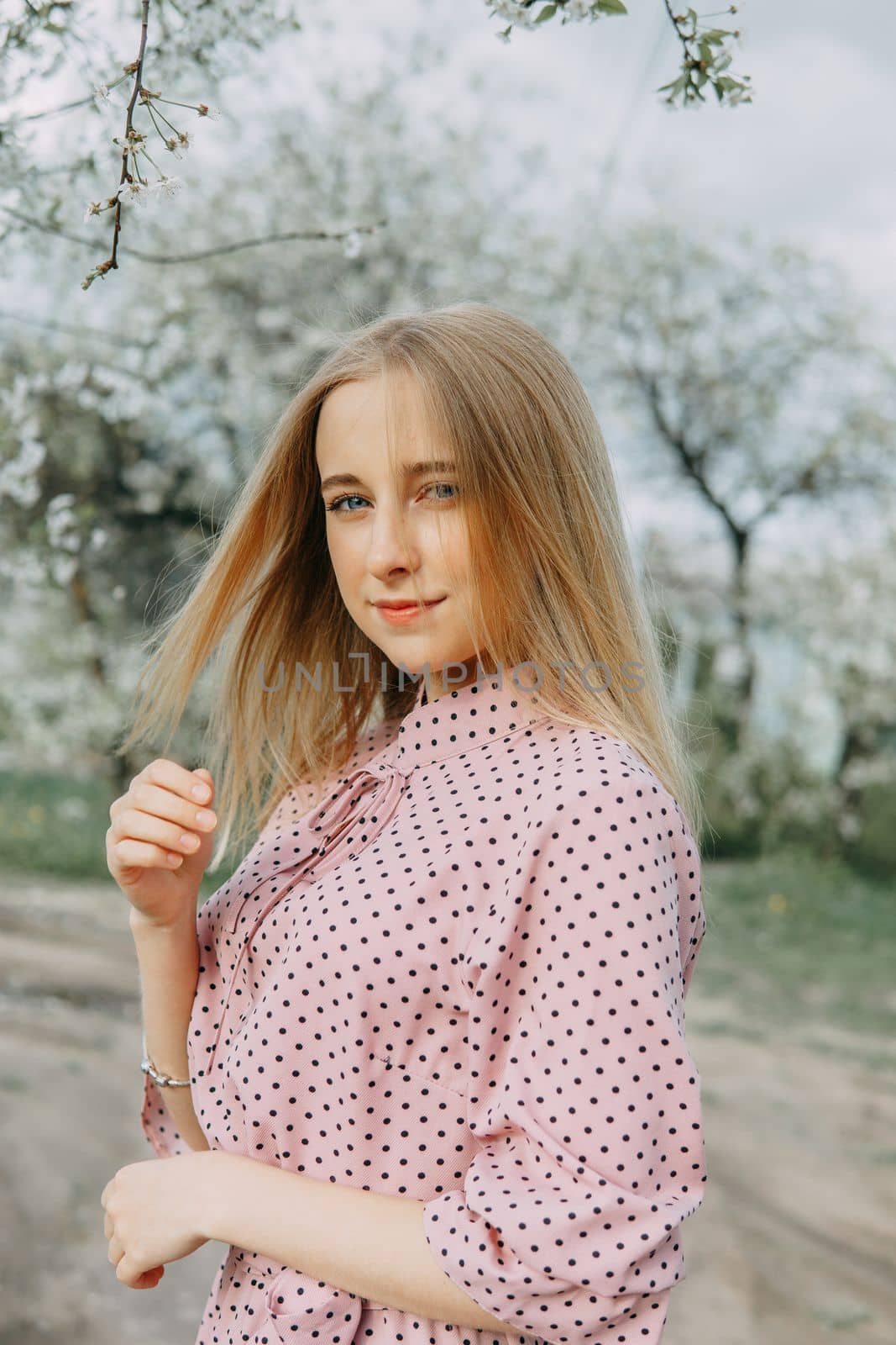 Blonde girl on a spring walk in the garden with cherry blossoms. Female portrait, close-up. A girl in a pink polka dot dress