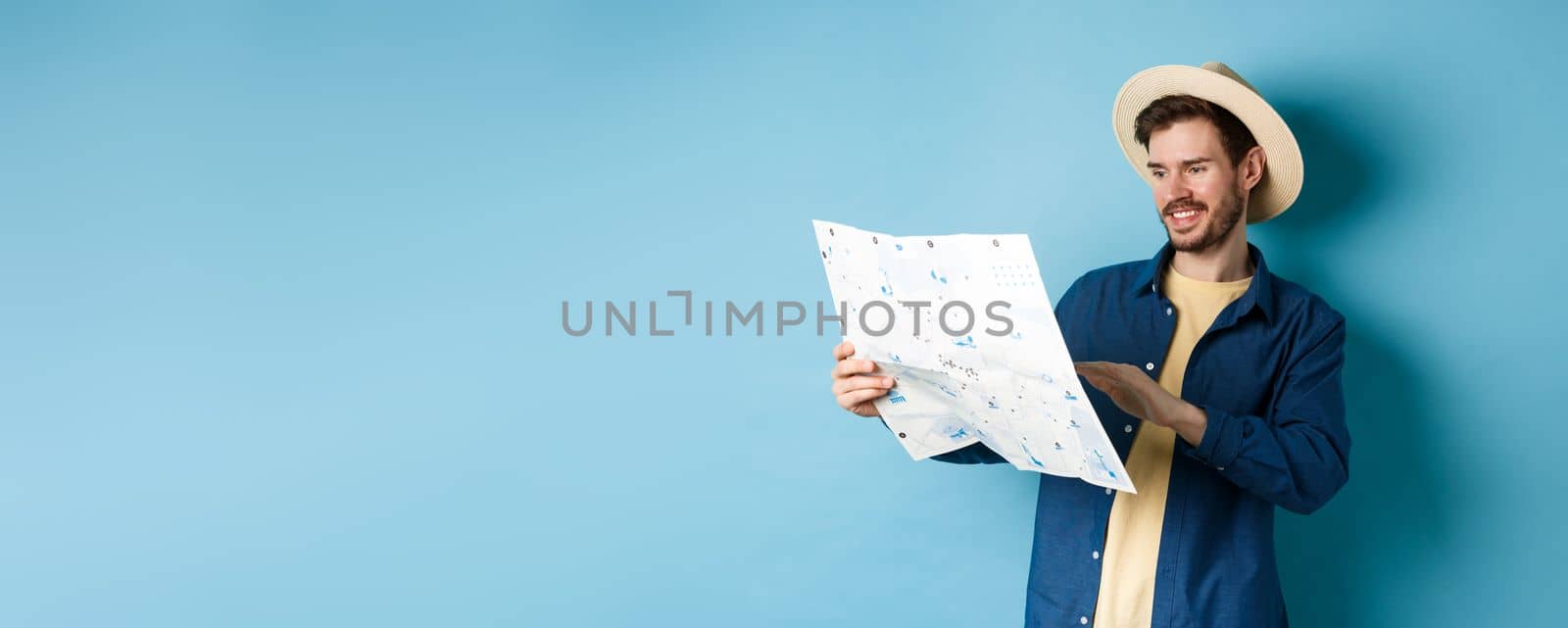 Handsome smiling tourist in straw hat looking at map, choosing travel road, planning a vacation, standing on blue background.