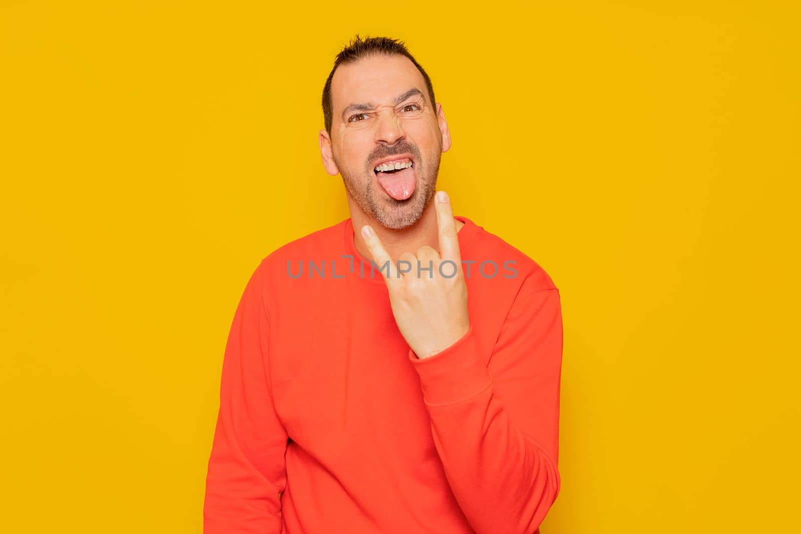 Bearded hispanic man wearing red pullover isolated over yellow background showing rock gesture with fingers and sticking out tongue