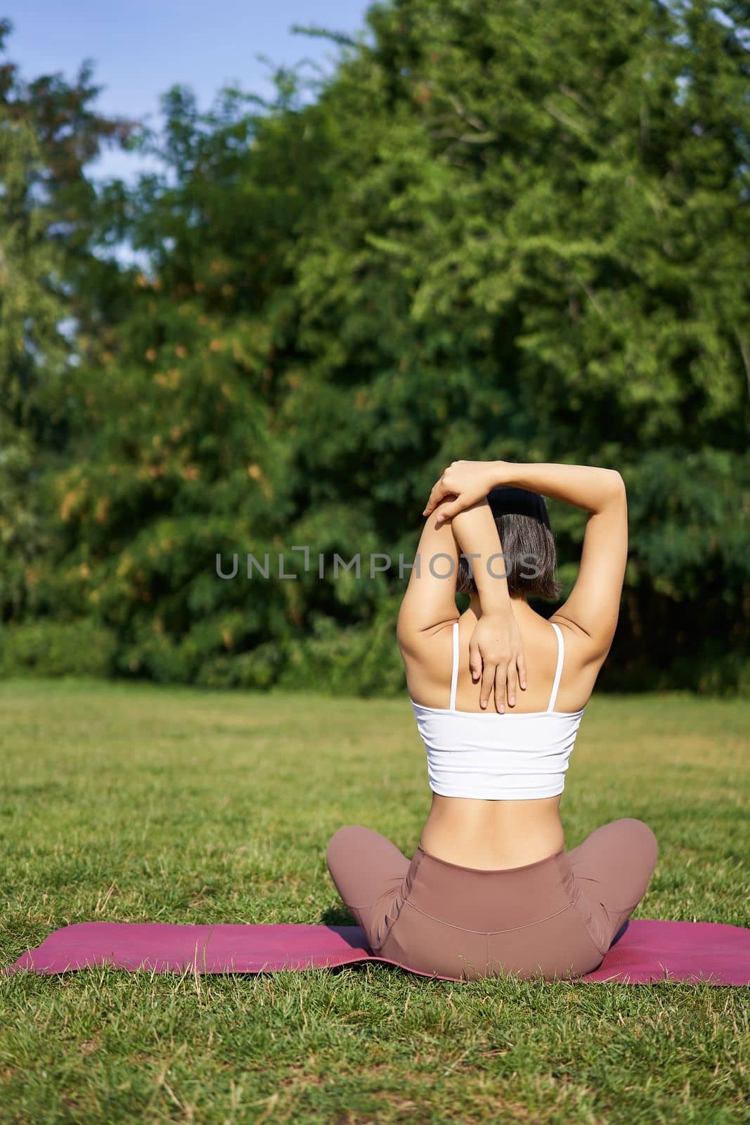 Rear view of sportswoman in middle of workout, stretching her amrs behind back, sitting on rubber yoga mat on lawn in park.