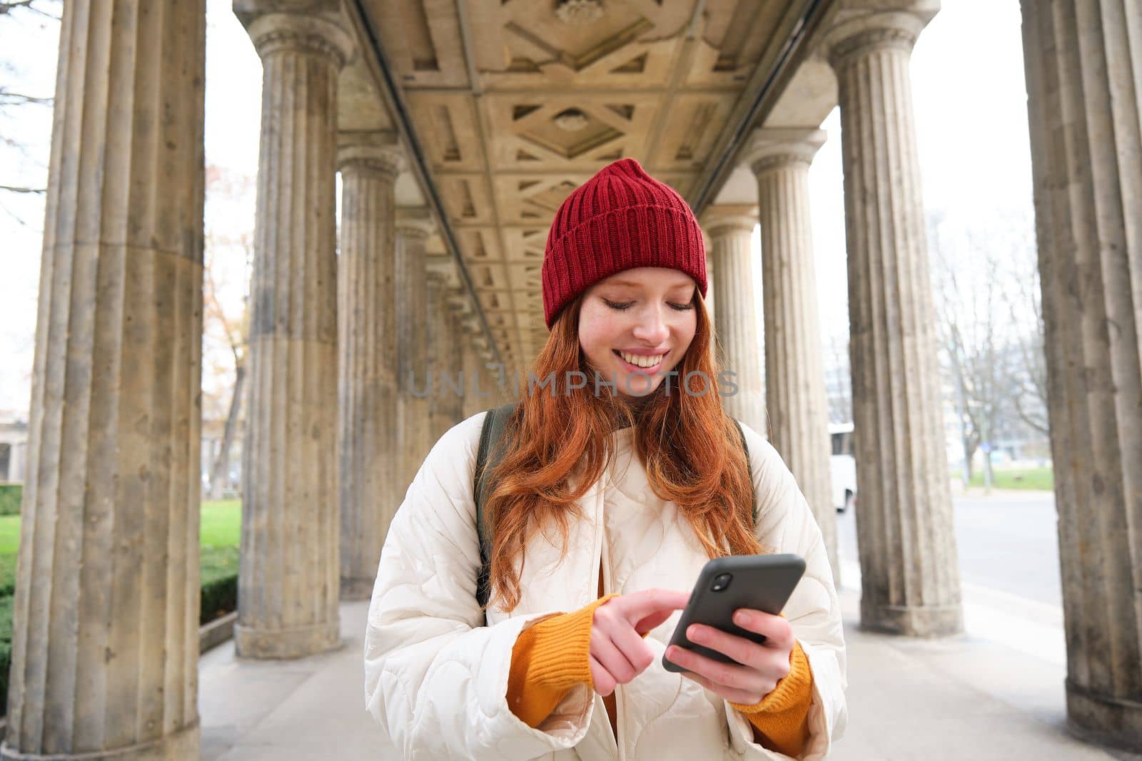 Mobile broadband and people. Smiling redhead 20s girl with backpack, uses smartphone on street, holds mobile phone and looks at application.