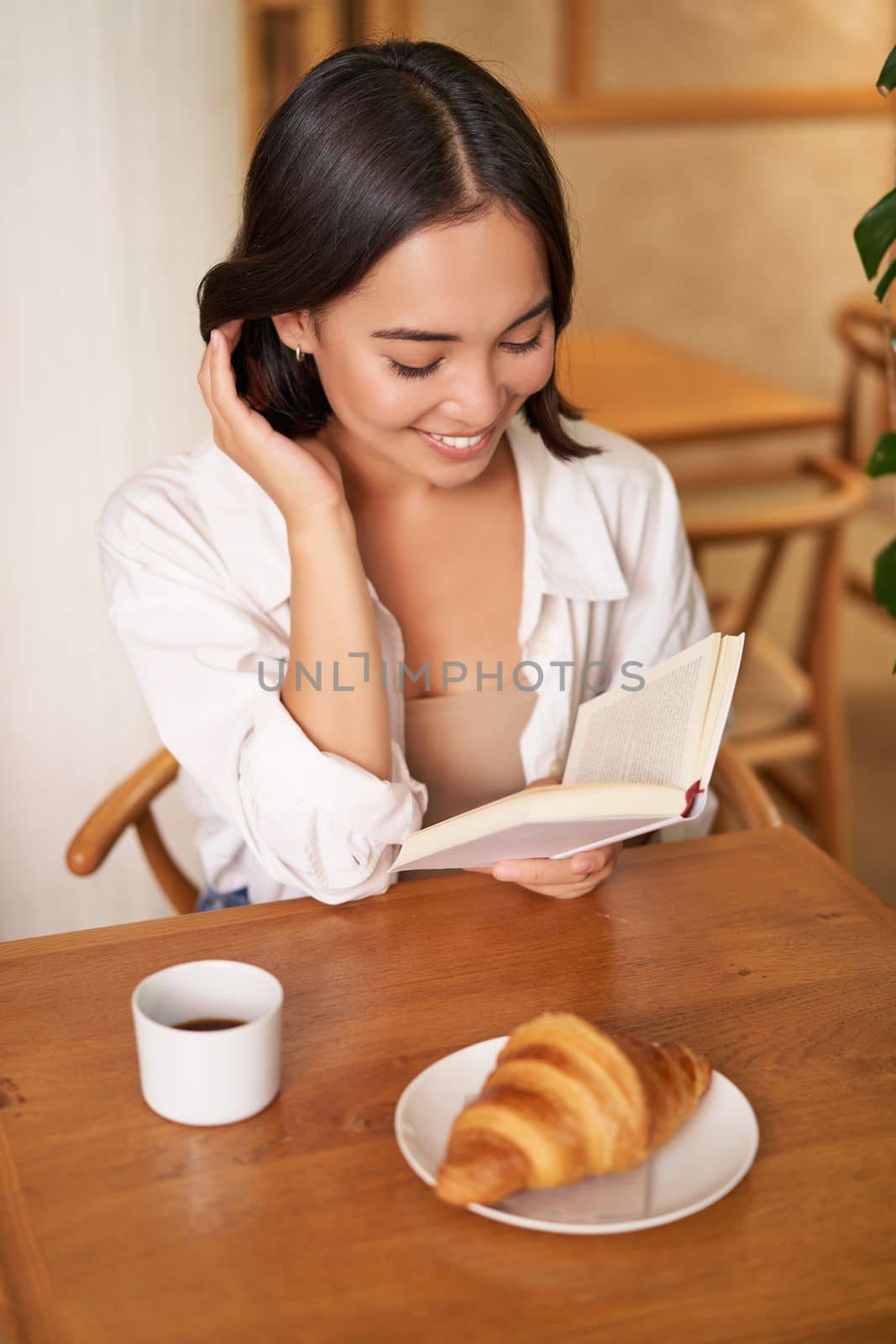 Vertical shot of happy young asian woman enjoys reading, sitting with book in cafe, drinking coffee and eating croissant.