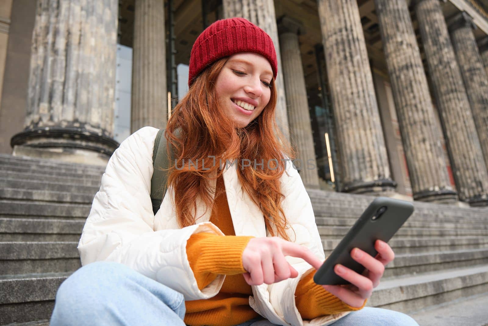 Stylish young redhead woman, talking on mobile phone app, using social media application, looking for something online on smartphone, sits on stairs outdoors by Benzoix