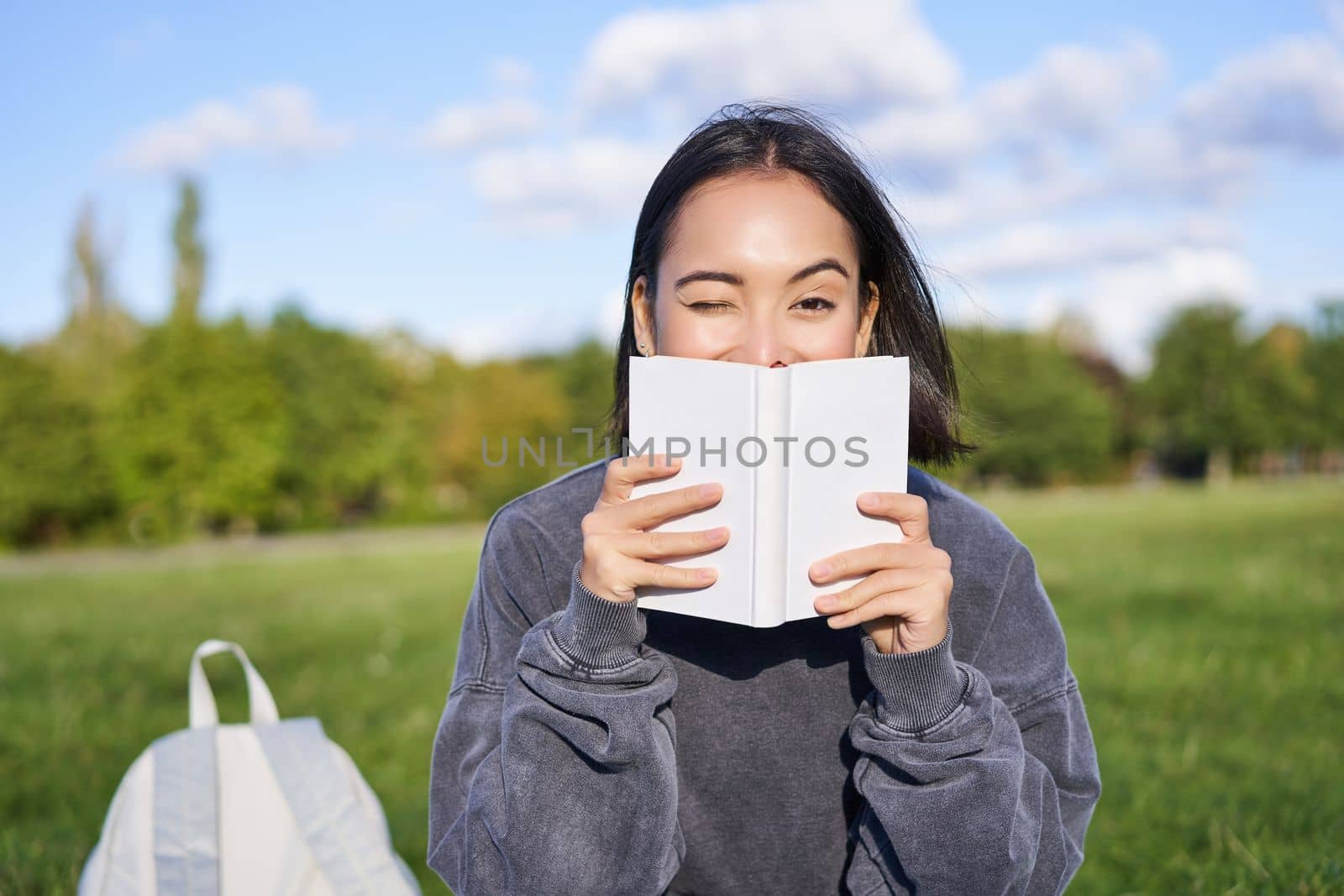 Beautiful asian girl sitting in park on grass, reading and smiling. Woman with book enjoying sunny day outdoors.