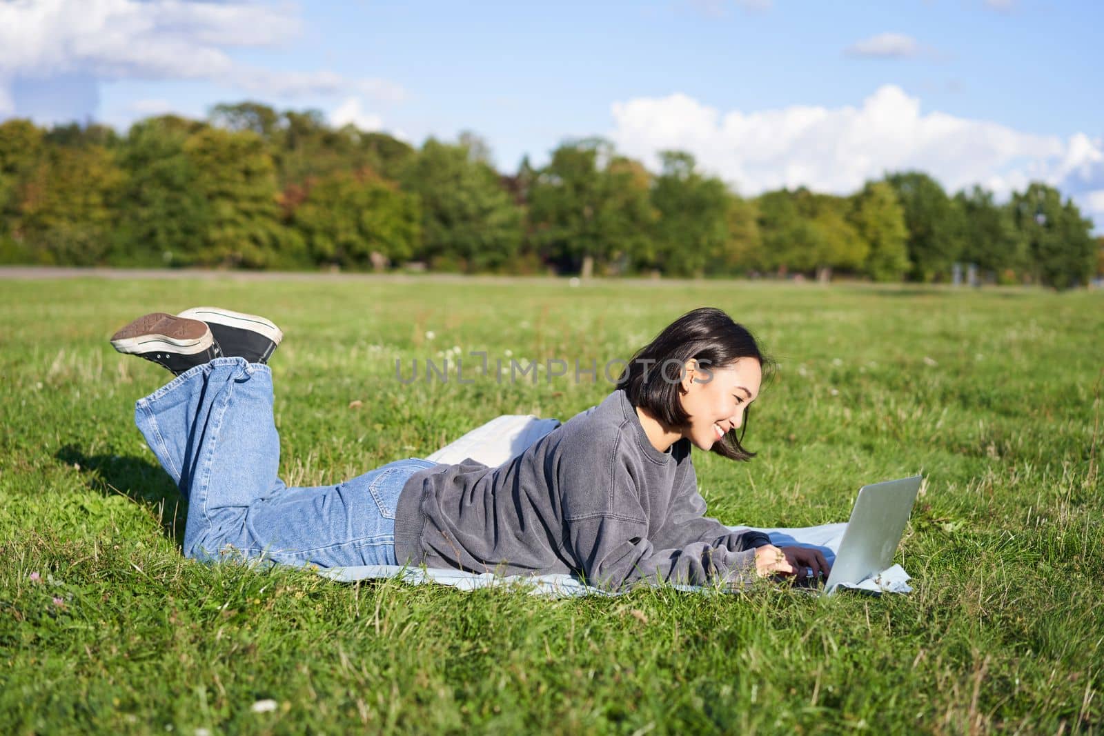 Portrait of young happy asian girl lying on blanket in park, watching videos and browsing internet on her laptop.