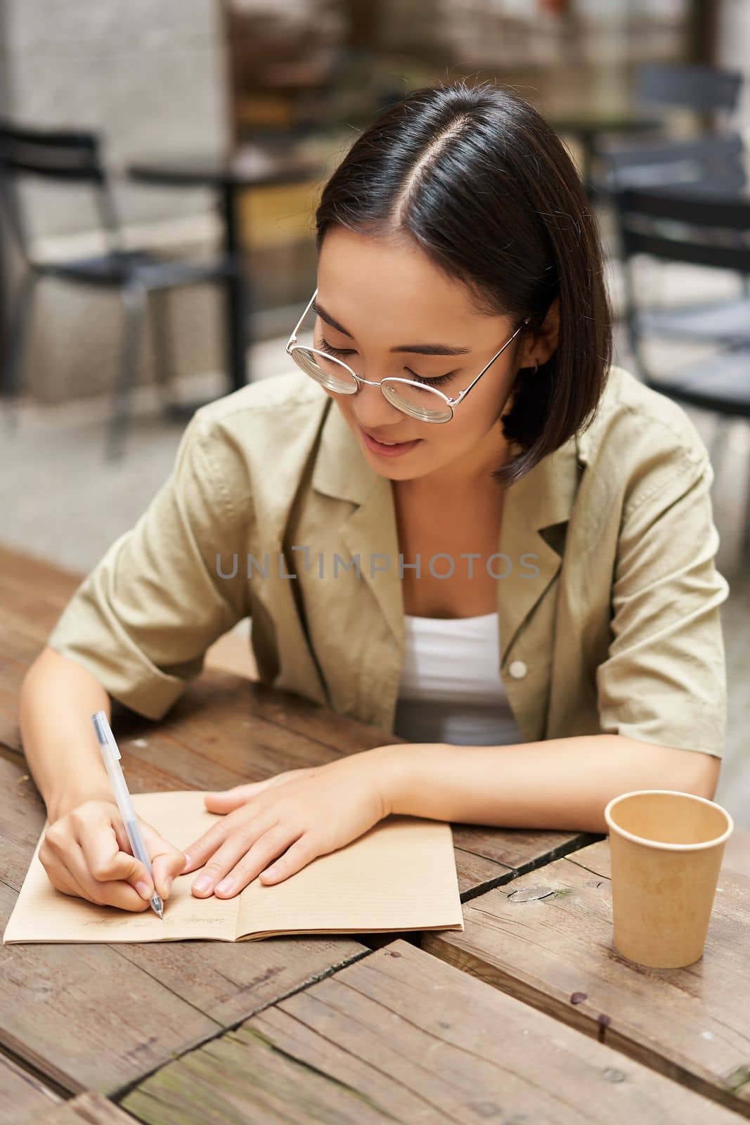 Vertical shot of young asian woman doing homework, making notes, writing something down, sitting in an outdoors cafe and drinking coffee.