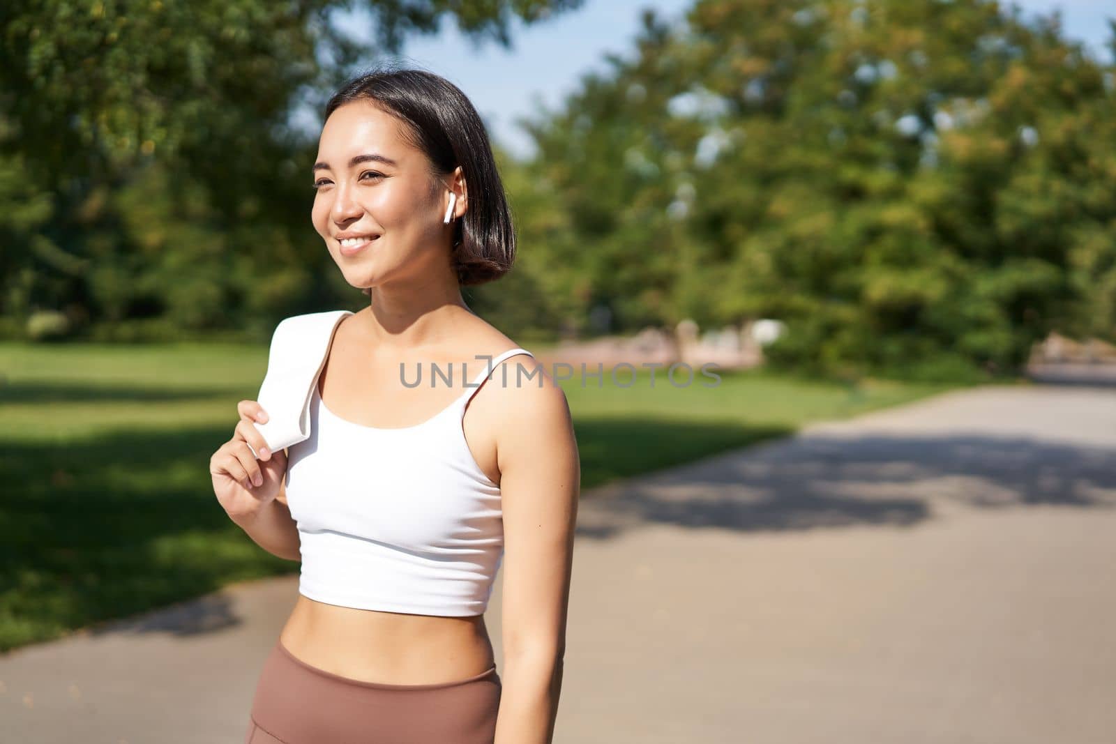 Smiling asian fitness girl holding towel on shoulder, workout in park, sweating after training exercises outdoors.